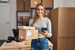 Young blonde woman ecommerce business worker using smartphone holding packages at office