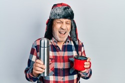 Handsome mature handyman wearing winter hat with ear flaps drinking hot coffee from thermo smiling and laughing hard out loud because funny crazy joke. 