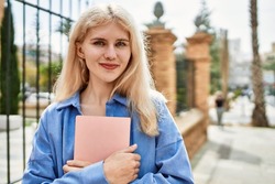 Beautiful blonde young woman smiling happy holding book at university campus on a sunny day