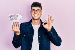 Young hispanic man holding egyptian pounds banknotes doing ok sign with fingers, smiling friendly gesturing excellent symbol 