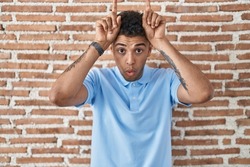 Brazilian young man standing over brick wall doing funny gesture with finger over head as bull horns 