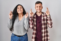 Young hispanic couple standing over white background gesturing finger crossed smiling with hope and eyes closed. luck and superstitious concept. 