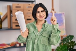 Young asian woman holding paper at art studio smiling with an idea or question pointing finger with happy face, number one 