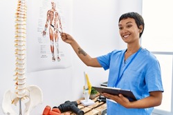 Young hispanic woman wearing physiotherapist uniform pointing to vertebral column holding checklist at physiotherapy clinic
