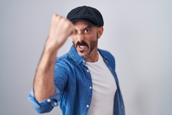 Hispanic man with beard standing over isolated background angry and mad raising fist frustrated and furious while shouting with anger. rage and aggressive concept. 