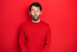 Handsome man with beard wearing casual red sweater making fish face with lips, crazy and comical gesture. funny expression. 