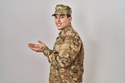 Young arab man wearing camouflage army uniform pointing aside with hands open palms showing copy space, presenting advertisement smiling excited happy 