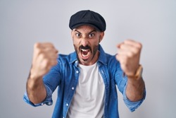 Hispanic man with beard standing over isolated background angry and mad raising fists frustrated and furious while shouting with anger. rage and aggressive concept. 