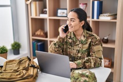 Young hispanic woman army soldier using laptop and talking on martphone at home