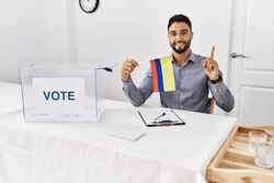 Young handsome man with beard at political campaign election holding colombia flag surprised with an idea or question pointing finger with happy face, number one 