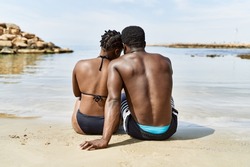 Young african american tourist couple on back view wearing swimwear sitting at the beach.
