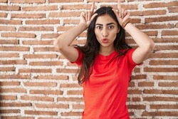 Young teenager girl standing over bricks wall doing funny gesture with finger over head as bull horns 