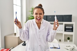 Young hispanic dentist woman holding ordinary toothbrush and electric toothbrush angry and mad screaming frustrated and furious, shouting with anger. rage and aggressive concept. 