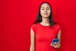 Young brazilian woman using smartphone over red background relaxed with serious expression on face. simple and natural looking at the camera. 