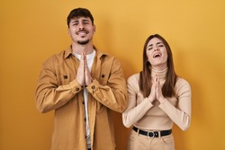 Young hispanic couple standing over yellow background begging and praying with hands together with hope expression on face very emotional and worried. begging. 