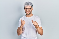 Young hispanic man with modern dyed hair wearing white t shirt and glasses pointing to the back behind with hand and thumbs up, smiling confident 