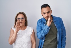 Hispanic mother and son standing together hand on mouth telling secret rumor, whispering malicious talk conversation 