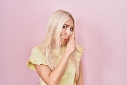 Caucasian woman standing over pink background hand on mouth telling secret rumor, whispering malicious talk conversation 