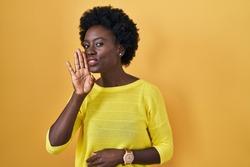 African young woman standing over yellow studio hand on mouth telling secret rumor, whispering malicious talk conversation 