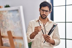 Young hispanic man artist holding paintbrushes with serious expression at art studio