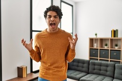 Young hispanic man wearing casual clothes standing at home crazy and mad shouting and yelling with aggressive expression and arms raised. frustration concept. 