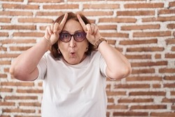 Senior woman with glasses standing over bricks wall doing funny gesture with finger over head as bull horns 