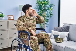 Arab man wearing camouflage army uniform sitting on wheelchair shouting and screaming loud to side with hand on mouth. communication concept. 