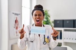 Young african american with braids wearing doctor uniform holding safety mask angry and mad screaming frustrated and furious, shouting with anger. rage and aggressive concept. 