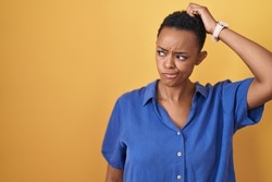 African american woman standing over yellow background confuse and wondering about question. uncertain with doubt, thinking with hand on head. pensive concept. 