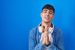 Young hispanic man standing over blue background begging and praying with hands together with hope expression on face very emotional and worried. begging. 