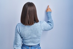 Young hispanic woman standing over blue background posing backwards pointing ahead with finger hand 