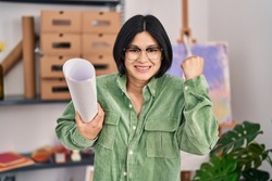 Young asian woman holding paper at art studio screaming proud, celebrating victory and success very excited with raised arm 