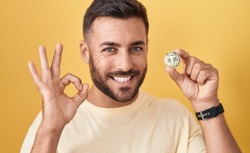 Handsome hispanic man holding tether cryptocurrency coin doing ok sign with fingers, smiling friendly gesturing excellent symbol 