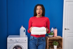 Young african american with braids holding clean laundry afraid and shocked with surprise and amazed expression, fear and excited face. 