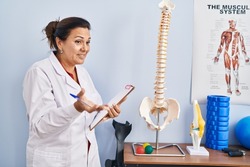 Middle age hispanic woman physiotherapist pointing to anatomical model of vertebral column at rehab clinic
