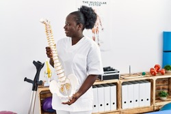 Young african american woman wearing physiotherapist uniform holding anatomical model of vertebral column at clinic