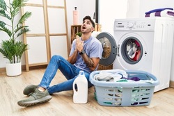 Young hispanic man putting dirty laundry into washing machine begging and praying with hands together with hope expression on face very emotional and worried. begging. 