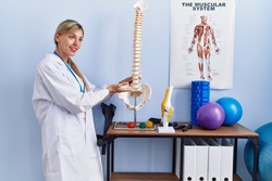 Young blonde woman wearing physiotherpist uniform pointing to anatomical model of vertebral column at physiotherapy clinic