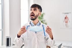 Young hispanic doctor man with beard holding safety mask angry and mad screaming frustrated and furious, shouting with anger. rage and aggressive concept. 