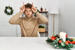 Young handsome man with beard sitting on the table by christmas decoration doing funny gesture with finger over head as bull horns 