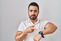 Young hispanic man with beard wearing casual clothes over white background in hurry pointing to watch time, impatience, upset and angry for deadline delay 