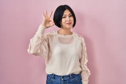 Young asian woman standing over pink background smiling positive doing ok sign with hand and fingers. successful expression. 