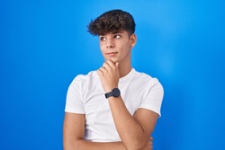 Hispanic teenager standing over blue background with hand on chin thinking about question, pensive expression. smiling with thoughtful face. doubt concept. 