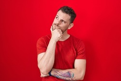 Young hispanic man standing over red background with hand on chin thinking about question, pensive expression. smiling with thoughtful face. doubt concept. 