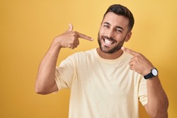 Handsome hispanic man standing over yellow background smiling cheerful showing and pointing with fingers teeth and mouth. dental health concept. 