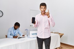 Young hispanic men at political campaign election holding smartphone surprised with an idea or question pointing finger with happy face, number one 