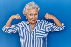 Senior woman with grey hair standing over blue background showing arms muscles smiling proud. fitness concept. 