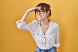 Young beautiful woman wearing casual shirt over yellow background very happy and smiling looking far away with hand over head. searching concept. 