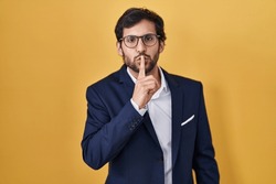 Handsome latin man standing over yellow background asking to be quiet with finger on lips. silence and secret concept. 