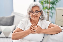 Middle age woman with hands on heart sitting on sofa at home